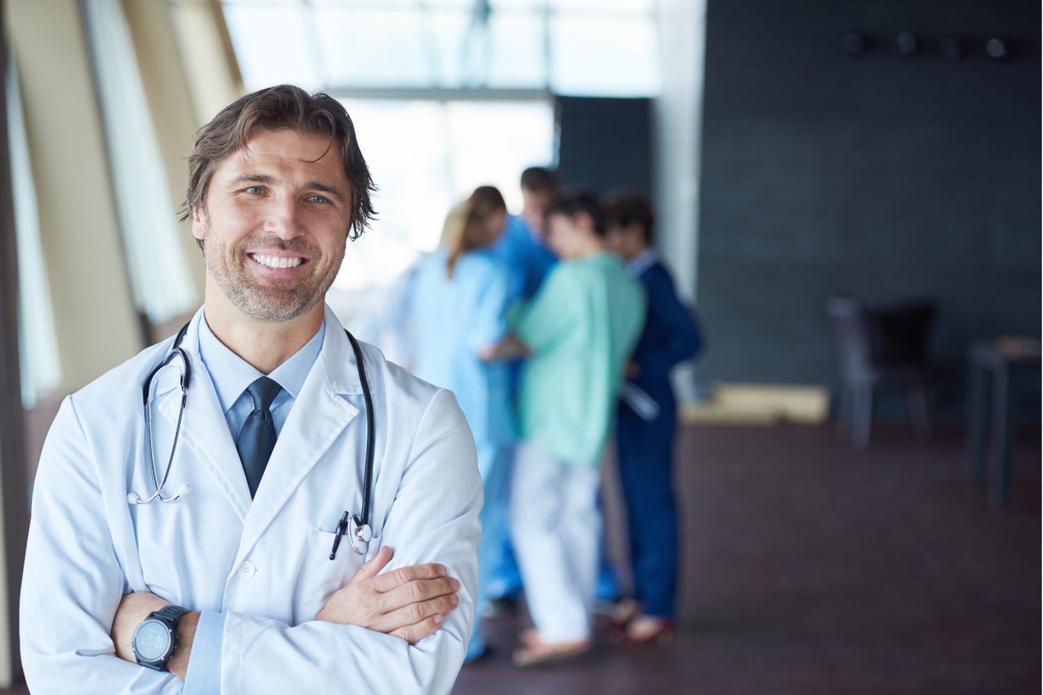 Male doctor stands, arms crossed, smiling beyond the camera. A group of nurses gather behind him.