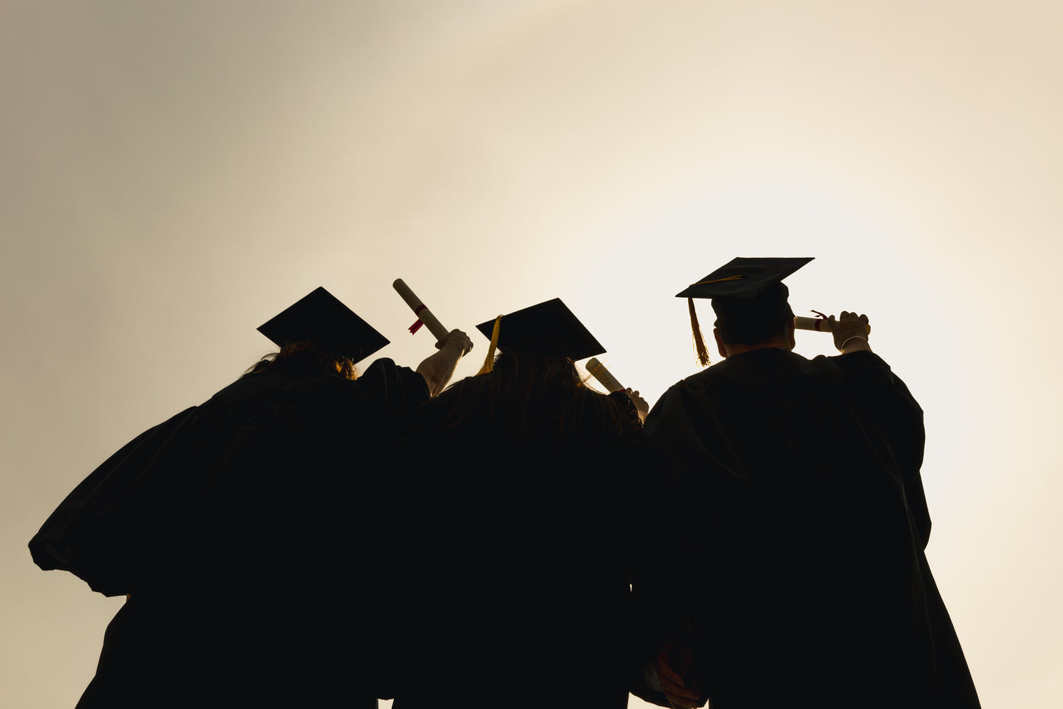 Silhouetted group of graduates hold their diplomas towards the sky.