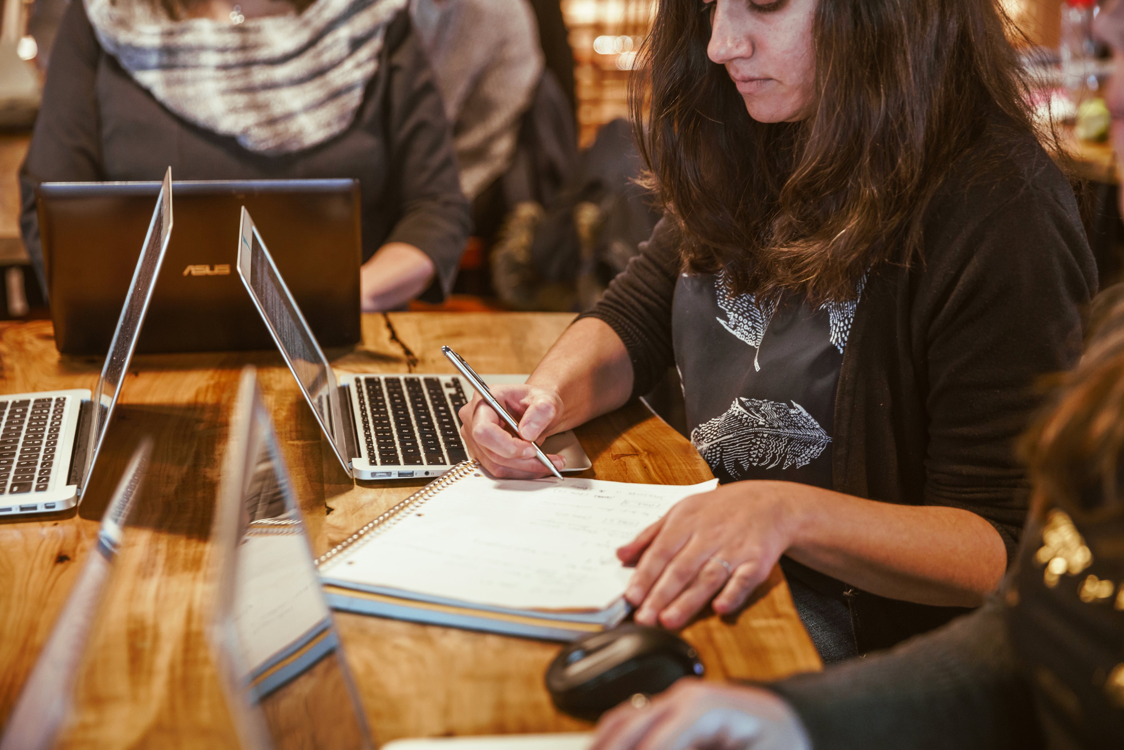 Group of young women at a table using laptops and writing in notebooks.