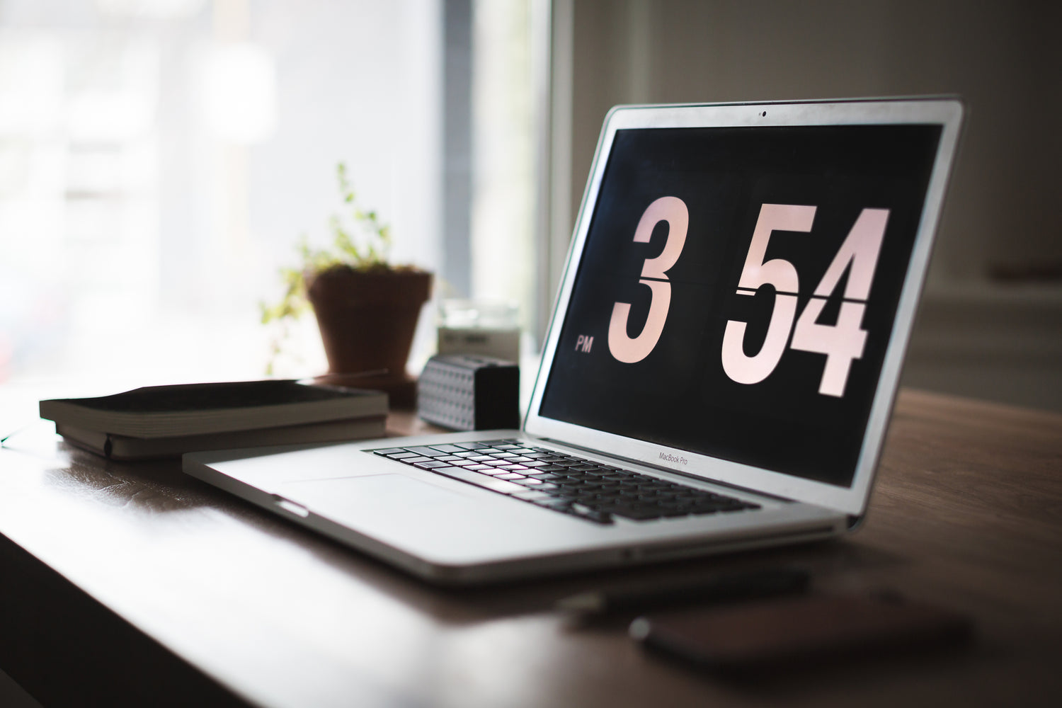 Open laptop on desk with a flip-clock screensaver reading 3:54 PM.