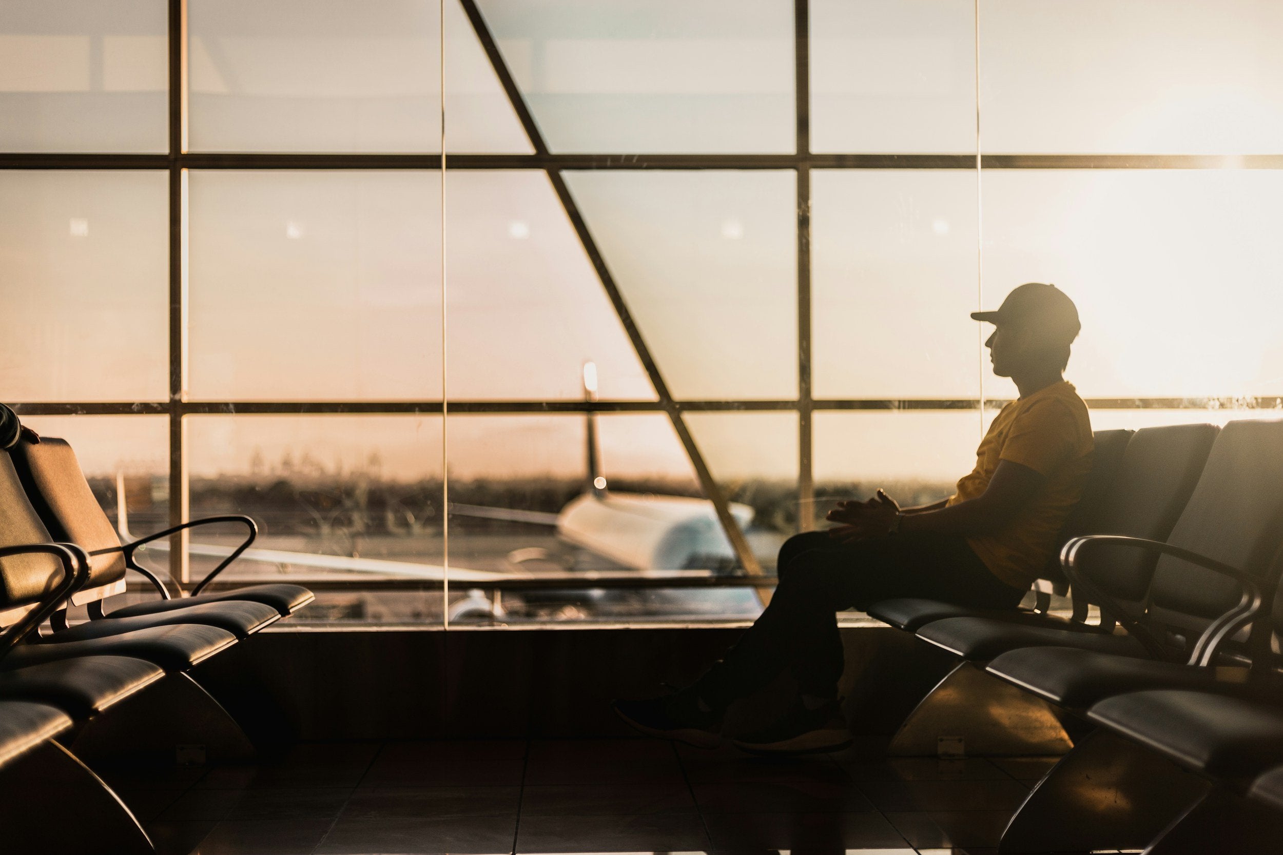 Man sitting at a gate in front of large wall of windows. Airplane is seen on tarmac in the background.